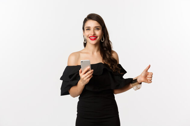 年轻网上购物概念穿着时髦的黑色连衣裙 化妆 竖起大拇指 使用手机应用程序 白色背景的女人女性Brunette女人
