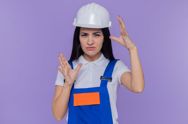 建筑身穿施工制服 头戴安全帽的年轻建筑女工人正注视着前方 双手站在紫色的墙壁上 专注于一个做工的姿势安全年轻女人
