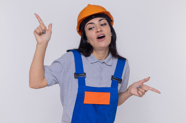 制服快乐和快乐的年轻建筑妇女穿着建筑制服和安全帽显示食指和双手站在白色的墙壁上建筑工人建筑手