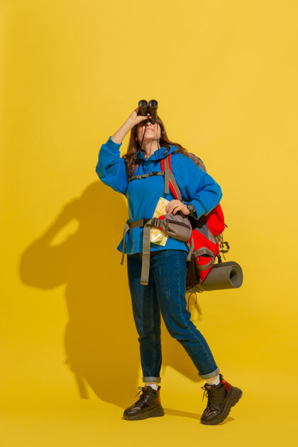 人黄色摄影棚墙上 一个带着包和望远镜的快乐的年轻旅游女孩的画像肖像女性假期