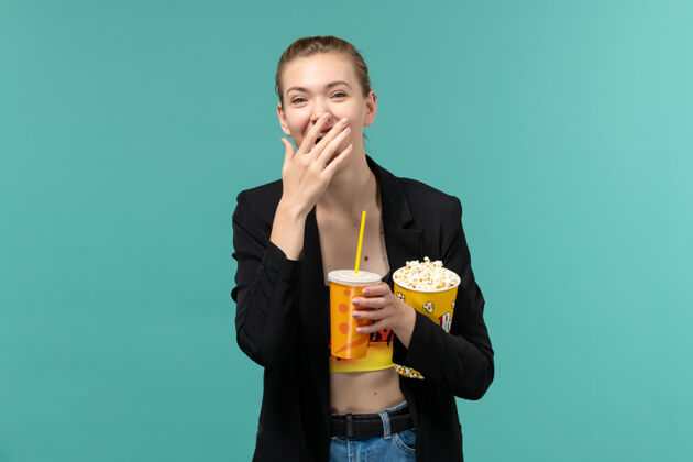 爆米花正面图：年轻女性拿着爆米花喝酒 在浅蓝色的表面上看电影电影剧院年轻的女性
