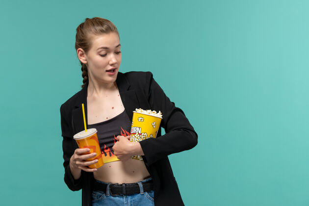 看正面图：年轻女性手持爆米花饮料 在浅蓝色的表面上看电影举行年轻的女性电影