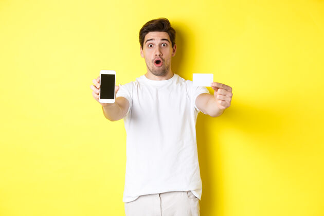 成人高加索帅哥展示智能手机屏幕和信用卡 手机银行和网上购物概念 黄色背景模特表情网购