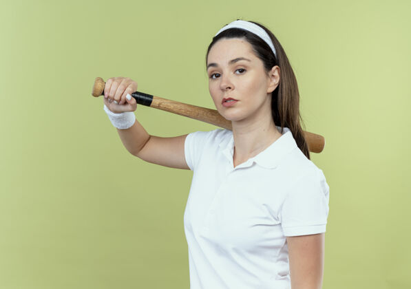 棒球戴着头巾 手持棒球棒 表情严肃自信的年轻健身女士站在轻盈的墙上站着轻女人