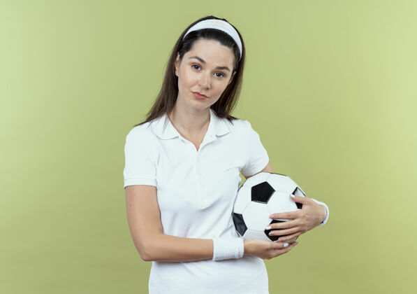 头带戴着头巾的年轻健身女士 手持足球 自信地站在轻盈的墙壁上立场健身信心