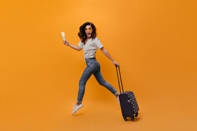 衬衫穿着牛仔裤的漂亮女孩在橙色背景上跳跃拿着票和手提箱的女人的全身画像头发包休闲