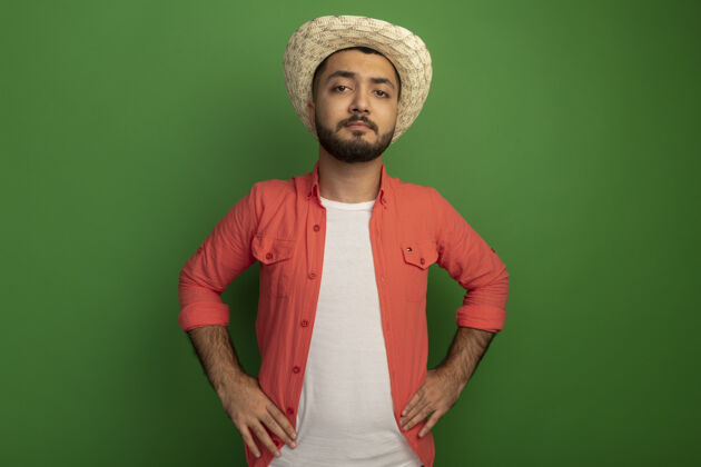 自信身穿橘色衬衫 头戴夏帽 留着胡子的年轻人站在绿色的墙上 神情严肃自信胡须夏天帽子