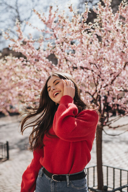 花园穿着红色毛衣的女士在樱花的背景下笑着玩弄着头发穿着开衫和牛仔裤的快乐女人在外面摆姿势自然衣服时尚