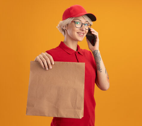 自信身穿红色制服 戴着帽子 戴着眼镜 拿着纸包 拿着手机 站在橘色的墙上自信地微笑着的年轻送货员眼镜站着拿着