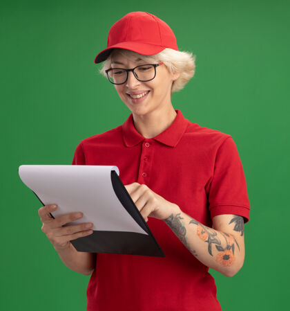 微笑身穿红色制服 戴着帽子 戴着眼镜的年轻女送货员站在绿色的墙上 脸上挂着空白的纸片 面带微笑地看着纸片制服站着脸