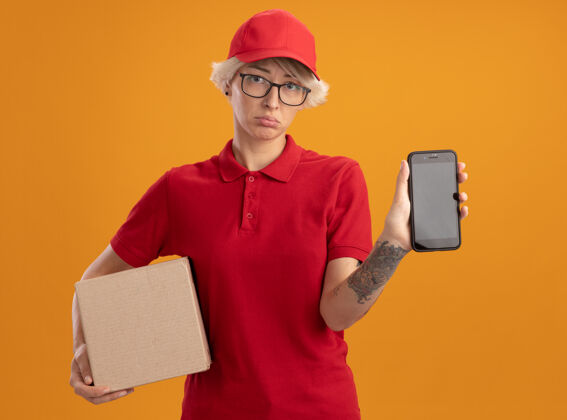 显示身穿红色制服 戴着帽子 戴着眼镜的年轻送货员拿着一个纸板箱 手里拿着一部智能手机 站在橙色的墙上 脸上带着悲伤的表情眼镜帽子穿