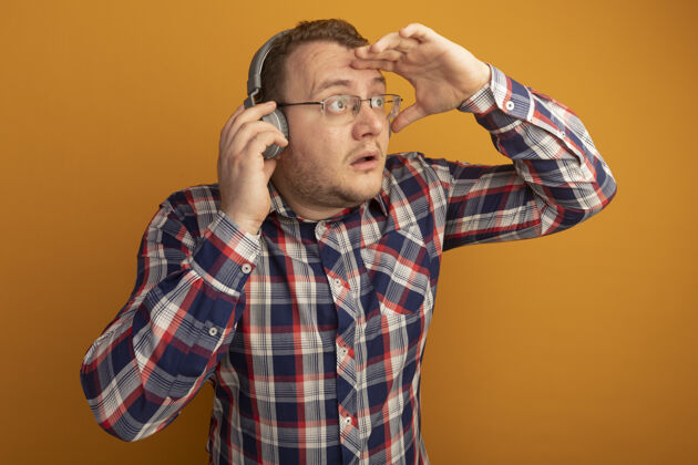 检查戴着眼镜和格子衬衫的男人戴着耳机望着远方 手举着头站在橙色的墙上耳机手离开