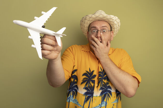 穿戴着橘色衬衫戴着夏帽戴着玩具飞机戴着眼镜的男人被吓得捂着嘴站在绿色的墙上站着拿着飞机