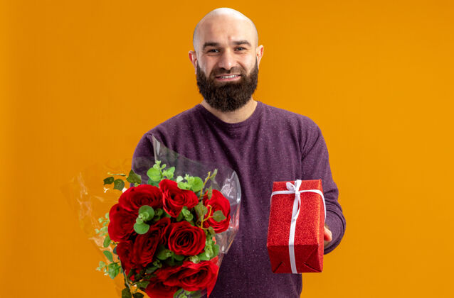 概念年轻的胡须男子手持一束红玫瑰和一份礼物微笑自信的情人节概念站在橙色的墙上花束微笑天