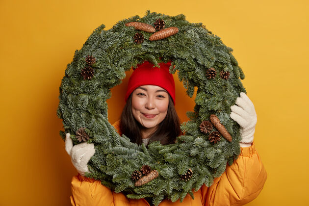 举行漂亮的亚洲女人戴着暖和的头套和手套 透过手工制作的传统圣诞花环 心情愉快 期待着寒假 隔离在黄色的工作室墙上乐观人类表情