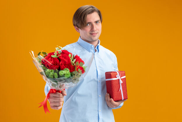 立场身穿蓝衣的男子手持一束红玫瑰 站在橙色的墙上 向大家展示微笑自信的情人节概念花束情人节玫瑰