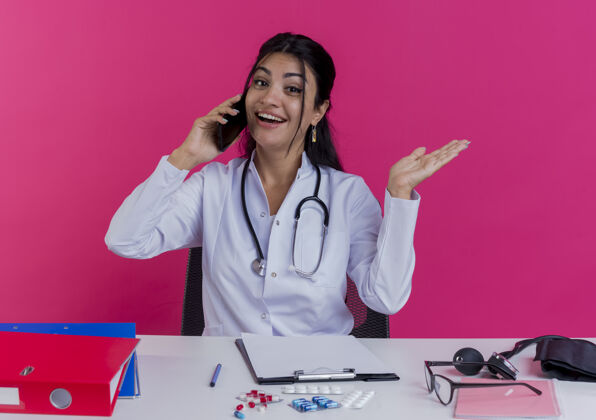 医生快乐的年轻女医生穿着医用长袍和听诊器坐在办公桌旁 手里拿着医疗工具在讲电话 粉红色的墙上空手孤立着穿欢乐谈话