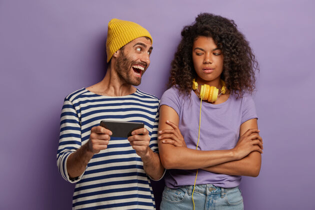 条纹无聊不高兴不满意的非洲裔美国少女双手交叉 看着朋友如何在现代智能手机上玩电子游戏 对新的应用程序赞不绝口T恤青少年消极