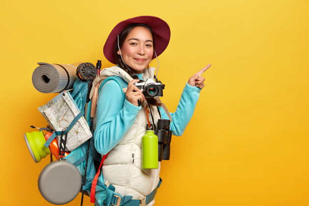 冒险活跃的女性背包客将食指指向旁边的复印空间 手持复古相机 拍照 携带背包 望远镜和保温瓶 穿着休闲服携带年份女人