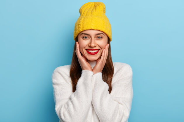 欢呼漂亮的女人穿红色唇膏 双手紧贴脸颊 戴黄色帽子和白色卡西米尔毛衣 对着蓝色的墙壁摆姿势舒适头口红
