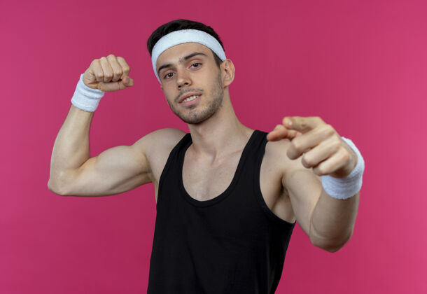 站着戴着头巾的年轻运动型男子自信地举起拳头 用食指指着镜头 越过粉色男人指向二头肌