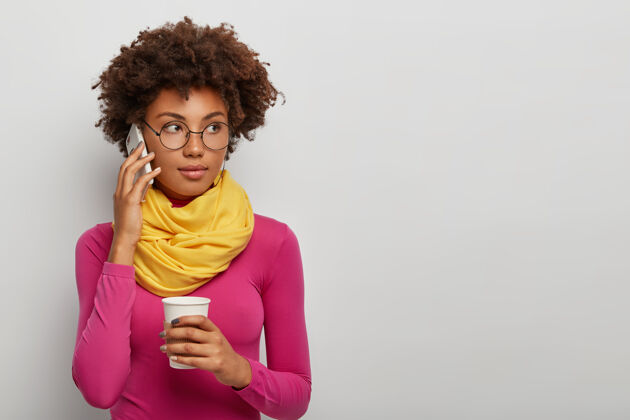 眼镜体贴的卷发女人通过手机通话 喝咖啡 享受交谈 戴眼镜 粉色高领毛衣配黄色围巾 在白色背景下摆姿势女性时尚肖像