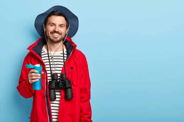 微笑高兴的男游客拿着蓝色的保温瓶 在旅游中喝茶 探索新的地方 穿着休闲服 带着望远镜 靠着蓝色的墙站着抱热水瓶男性