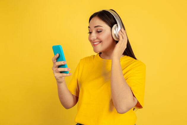 职业用无线耳机和手机听音乐黄色墙壁上的白种女人休闲的漂亮深色模特人类情感的概念 面部表情 销售 广告 广告空间快递现代年轻