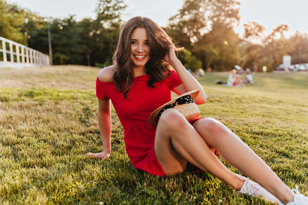 减肥美丽的女孩 羞涩的微笑 坐在公园的地上阳光明媚的日子里 一个穿着红裙子 幽默的黑发女人在草地上摆姿势年轻放松户外