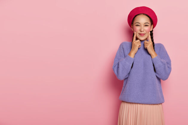 化妆积极的年轻女子用前指捂住两颊 精神抖擞 头戴红色贝雷帽 身穿紫色毛衣和裙子 站在粉红色的墙上室内女性感觉