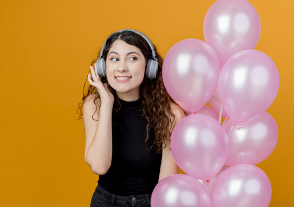 举行年轻漂亮的女人 卷发 戴着耳机 听音乐 手持一束气球 站在橘色的墙上 快乐快乐的生日派对气球欢呼听