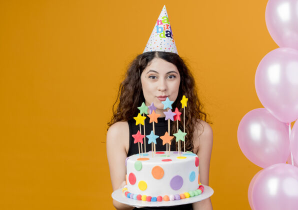 微笑一位年轻漂亮的女士 卷发 戴着节日帽 手里拿着生日蛋糕 微笑着 快乐地站在橙色的墙上 手里拿着气球气球蛋糕女人