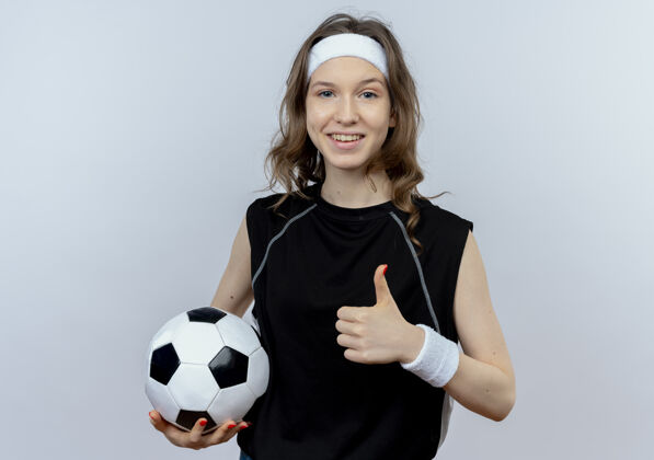 拇指身穿黑色运动服的年轻健身女孩 头箍举着足球 微笑着竖起大拇指站在白色的墙上微笑女孩头带
