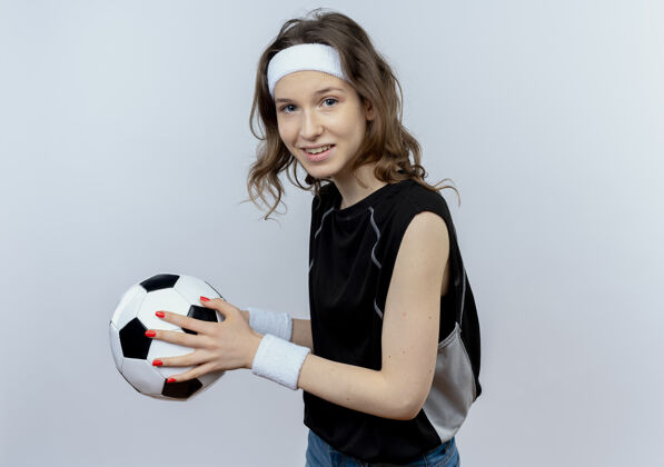站着身穿黑色运动服 头箍 手持足球 微笑着站在白墙上的年轻健身女孩年轻头带微笑
