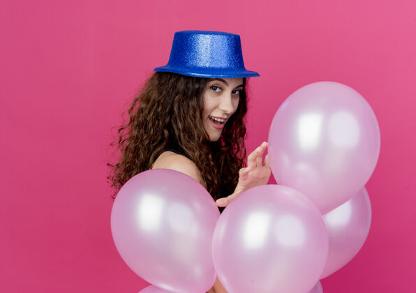 微笑一个年轻漂亮的女人 一头卷发 戴着节日礼帽 手里拿着一堆气球 站在粉色的墙上 开心而积极地笑着站着卷曲抱着
