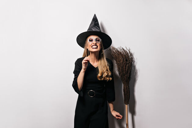成人快乐的女模特穿着有趣的万圣节服装摆姿势情绪激动的金发女孩戴着女巫帽站在白色的墙上年轻魔术师女巫
