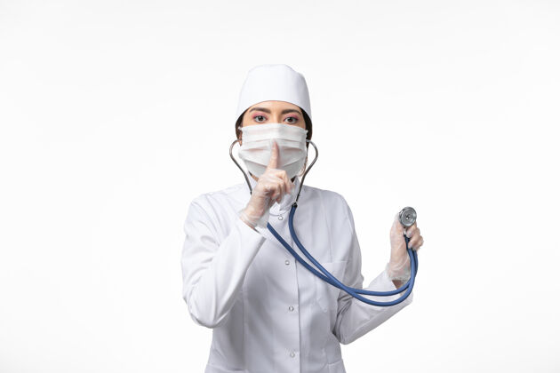 冠状病毒防护正面图女医生身穿白色无菌医疗服 戴口罩因冠状病毒-使用听诊器对白墙疾病冠状病毒-大流行性疾病冠状病毒预防视图使用