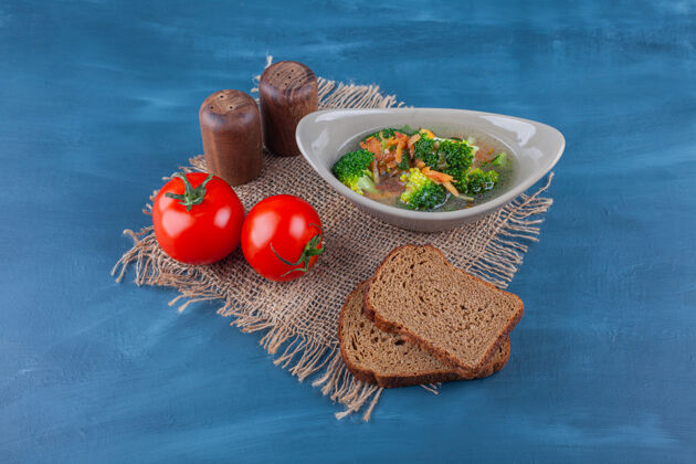 家禽一碗鸡汤 蔬菜和面包放在蓝色表面的粗麻布餐巾上配料番茄西兰花