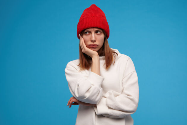孤独一个愁眉苦脸的女人 一头黑发的漂亮女孩穿着白毛衣 戴着红帽子把头放在一只手上感觉很无聊看着左边的复制空间 隔着蓝色的墙看起来情绪面部