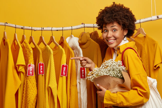 黄色快乐的年轻非洲裔美国妇女的水平镜头指向挂在铁轨上的时髦衣服 拎着包和漂亮的花束 有着牙齿般的微笑 隔离在黄色背景下设计师指标服装