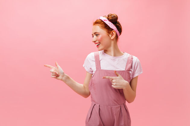 头带穿着粉色裙子和白色上衣的可爱女人在孤立的背景上展示手指在左边性感时尚模特
