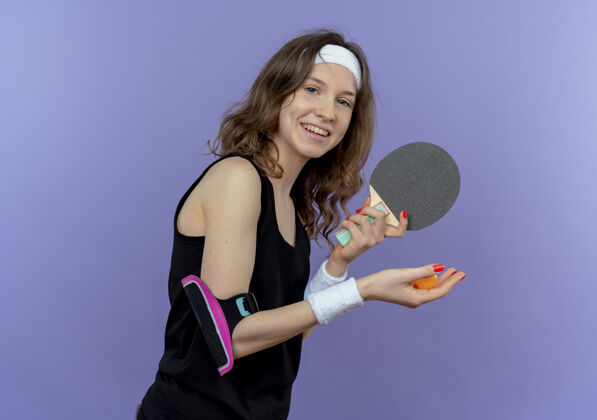准备身穿黑色运动服的年轻健身女孩 头上戴着头带 手里拿着球拍和乒乓球 准备好打乒乓球了 站在蓝色的墙上 她笑得很开心头带健身球