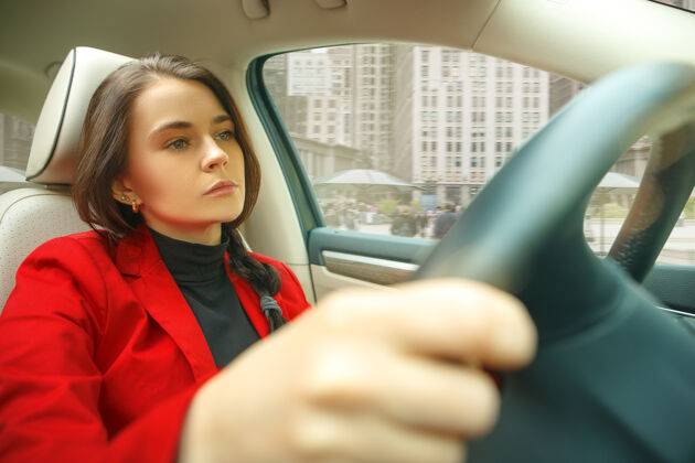 人在城市里开车年轻漂亮的女人开着车年轻漂亮的白人模特穿着优雅时尚的红色夹克坐在现代汽车内饰上女商人概念坐着开车内装