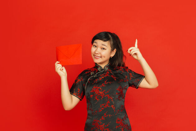 红色中国新年红色背景上孤立的亚洲少女肖像身着传统服装的女模特看起来很开心 微笑着指着红包庆祝 节日 情感装饰年祝福