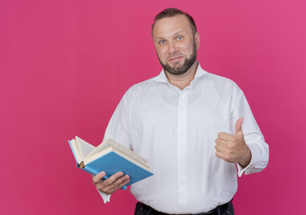 微笑留着胡子的男人穿着白衬衫 拿着打开的书微笑着竖起大拇指站在粉红色的墙上拇指抱着张开