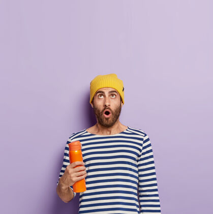 茶惊艳的年轻男模照片向上聚焦 手持橙色保温瓶 身穿条纹套头衫 头戴黄色帽子 有着惊艳的表情紫色难以置信室内