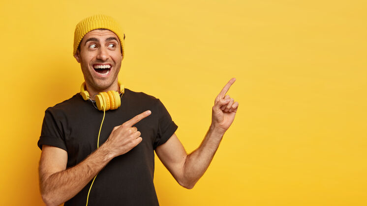 黑色横向拍摄的快乐欧洲男子用两个食指指着旁边 穿着时尚的黑黄相间的衣服 脖子上戴着现代耳机听歌头饰手指聆听