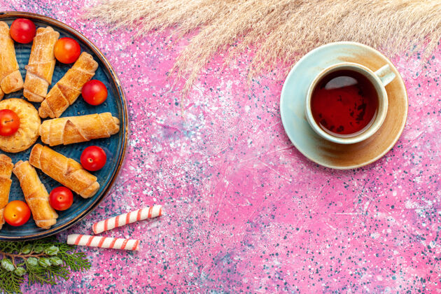 新鲜顶视图甜甜可口的百吉饼在托盘内与新鲜的酸李子和茶在淡粉色的桌子上杯子饼干糖