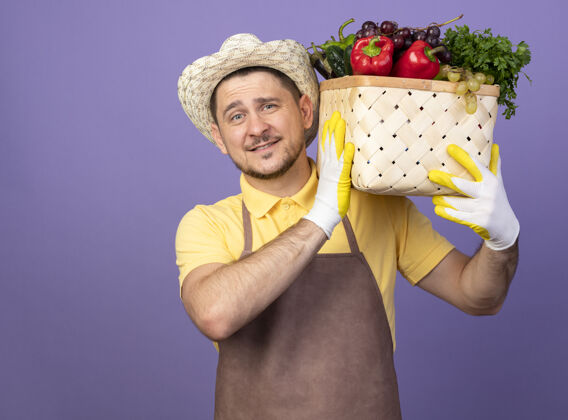 连身衣年轻的园丁穿着连体衣 戴着帽子 戴着工作手套 肩上扛着装满蔬菜的箱子 站在紫色的墙上 面带微笑地看着前方板条箱花园男人