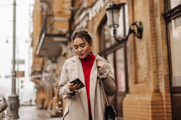 城市时尚的亚洲女士穿着米色外套 红色上衣和交叉体包 拿着智能手机在城市里走来走去自然头发街道